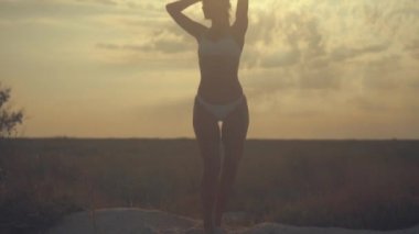 Genç güzel seksi tabaklanmış kız bikinili kumsalda gün batımında deniz kenarında dans kıvırcık saçlı. O obscurities gülümsedi, kendini ve yavaş hareket okşadı. 120fps