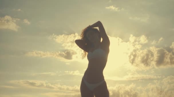 Krásná mladá sexy opálená dívka v bikinách s kudrnatými vlasy tančí na pláži při západu slunce u moře. Záhadně usmál, pohladil sama a. zpomaleně. 120fps — Stock video
