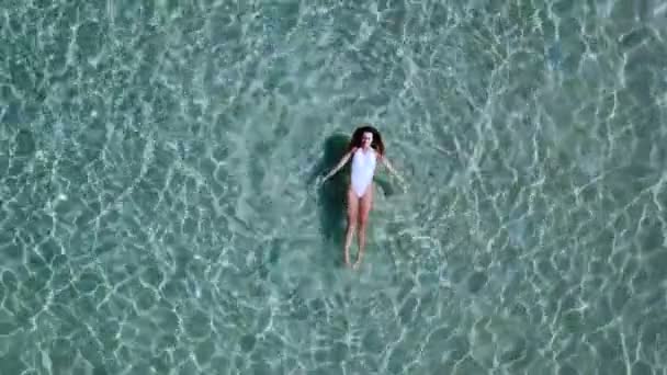 Vue aérienne. Belle jeune femme en bikini blanc flottant sur la surface de l'eau dans un océan de couleur turquoise cristalline — Video