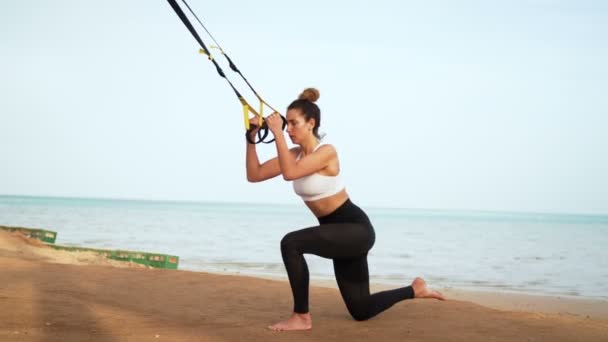 Atlética, mujer joven sexy, haciendo ejercicios con sistema de trx fitness, correas de suspensión TRX. En la playa, en verano, en los rayos del sol . — Vídeos de Stock