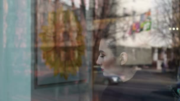 Una joven mirando por la ventana de una cafetería. Parece feliz. Vemos tráfico y gente en el reflejo de la ventana . — Vídeo de stock