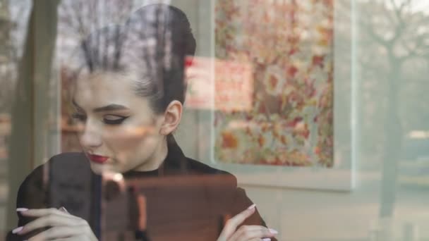 Ung kvinna tittar utanför genom fönstret på ett kafé. Hon ser glad. Vi se trafik och människor i reflektion av fönstret. — Stockvideo