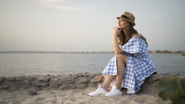 Belle femme en chapeau et robe est assis et regardant le coucher du soleil — Video