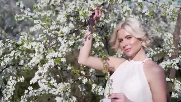 Adorable joven rubia con maquillaje es encantadora sonriendo y oliendo la flor blanca en el jardín floreciente. Moviéndose en el pelo del viento . — Vídeo de stock