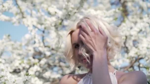 Adorable joven rubia con maquillaje es encantadora sonriendo y oliendo la flor blanca en el jardín floreciente. Moviéndose en el pelo del viento . — Vídeo de stock