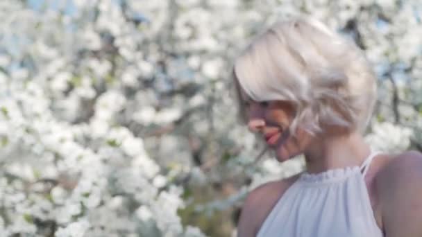 Roztomilá mladá blondýnka s make-up je milé, s úsměvem a vonící bílý květ v kvetoucí zahradě. Pohybující se ve vlasech vítr. — Stock video