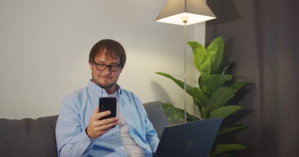Happy Young Man utiliza el ordenador portátil y smartpfone para el trabajo mientras está sentado en un sofá en casa . — Vídeos de Stock