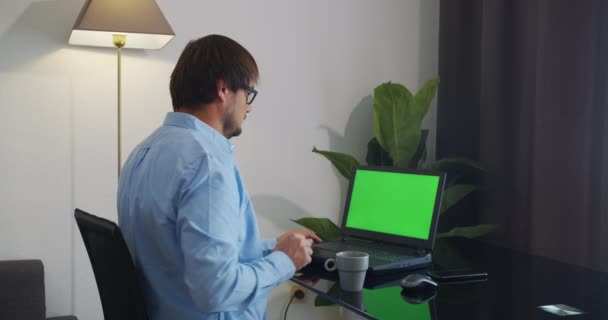 Человек дома сидит на диване и работает на зеленом макете экрана ноутбука. — стоковое видео