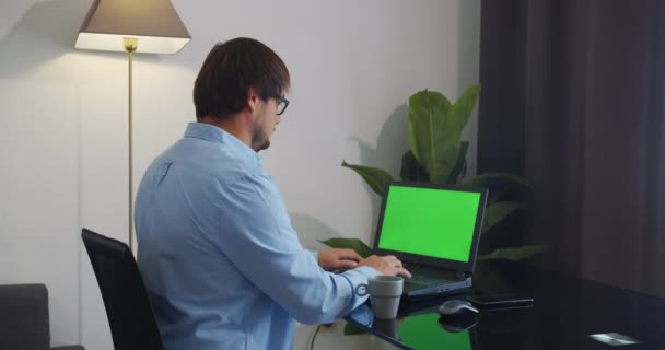 Homem em casa sentado em um sofá trabalhando em computador portátil tela verde mock-up. — Vídeo de Stock