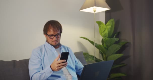 Jovem feliz usa laptop e smartpfone para o trabalho enquanto sentado em um sofá em casa . — Vídeo de Stock