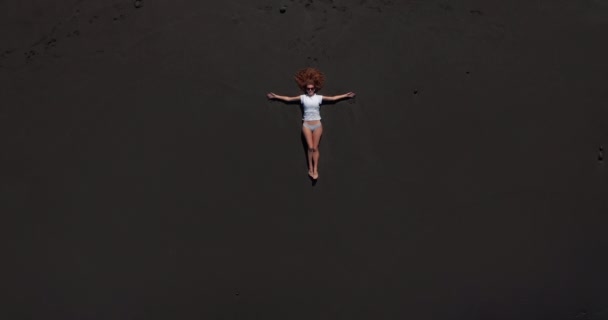 Aerial view. Beautiful woman lay down and rest on the black beach in tenerife while a long oceanic wave is coming. — Stock Video