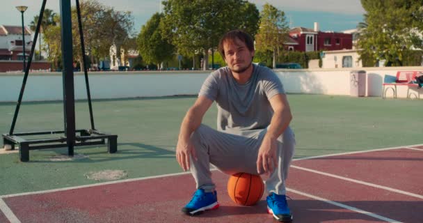 Баскетболіст сидить з м'ячем на баскетбольному майданчику. Молодий хлопець готується до баскетбольної гри . — стокове відео