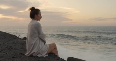 Romantik beyaz Avrupalı kız büyük bir kayanın üzerinde oturuyor. Sahilde gün batımında okyanus gelgiti.