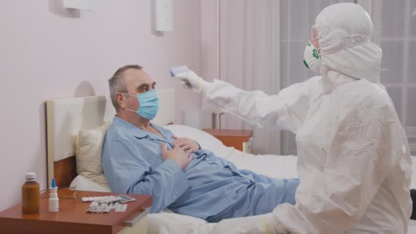 Temperatuurmeting van een zieke oudere man tijdens een epidemie. Infectiepreventie en beheersing van epidemieën. Beschermende kleding en masker. — Stockvideo