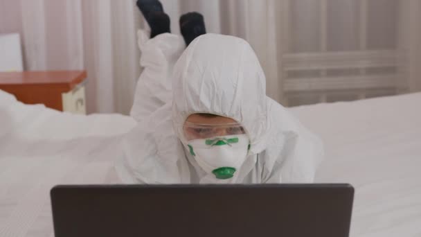 Ein Junge in Schutzanzug und Atemschutzmaske zu Hause am Computer, isoliert während einer Epidemie. — Stockvideo