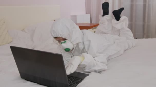 Ein Junge in Schutzanzug und Atemschutzmaske zu Hause am Computer, isoliert während einer Epidemie. — Stockvideo
