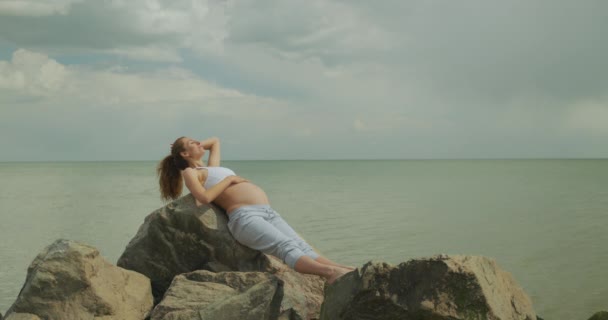Krásná těhotná žena na břehu moře, mladá žena čeká na své dítě, hladí ho po břiše. — Stock video