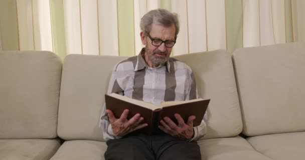 Άντρας συνταξιούχος διαβάζει ένα βιβλίο και αποκοιμιέται ενώ διαβάζει καθισμένος στον καναπέ. — Αρχείο Βίντεο