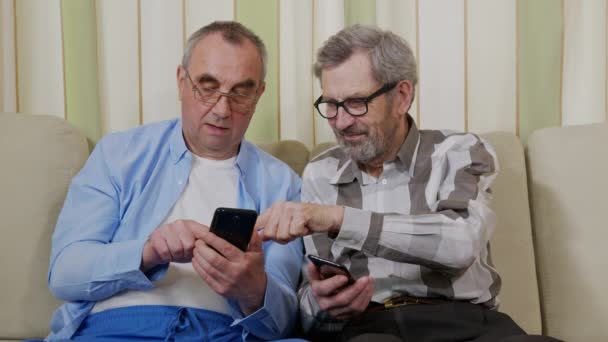 Dwóch starszych mężczyzn znających się na smartfonie i mówiących. Radosni emeryci rozmawiający na kanapie w domu opieki. — Wideo stockowe