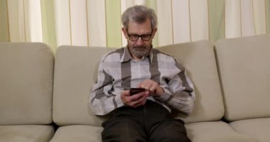 Yakın plan. Gözlüklü, telefonda konuşan emekli bir erkek. Evde akıllı telefon kullanan yaşlı bir emekli. Modern teknoloji, yaşam tarzı, yaşlanma, sakatlık.