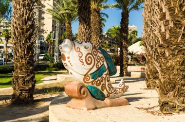 EILAT, ISRAEL - November 7, 2017: picturesque sculpture in form of fish in bright colors painted with floral patterns in beige and green tones standing among the palms. In background, urban buildings, Eilat, Israel  clipart