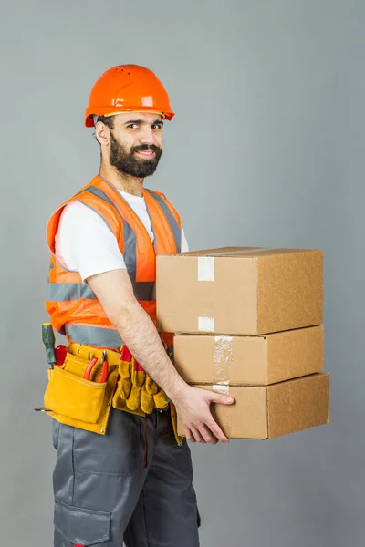Um construtor de homens com um capacete laranja com uma caixa de papelão nas mãos. Em um fundo cinza — Fotografia de Stock