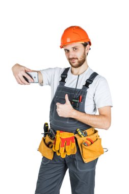 Bir işaret gösteren ve telefon resim alma genç inşaat işçisi gülümseyerek. Onarım ve inşaat. Beyaz arka plan üzerinde izole.
