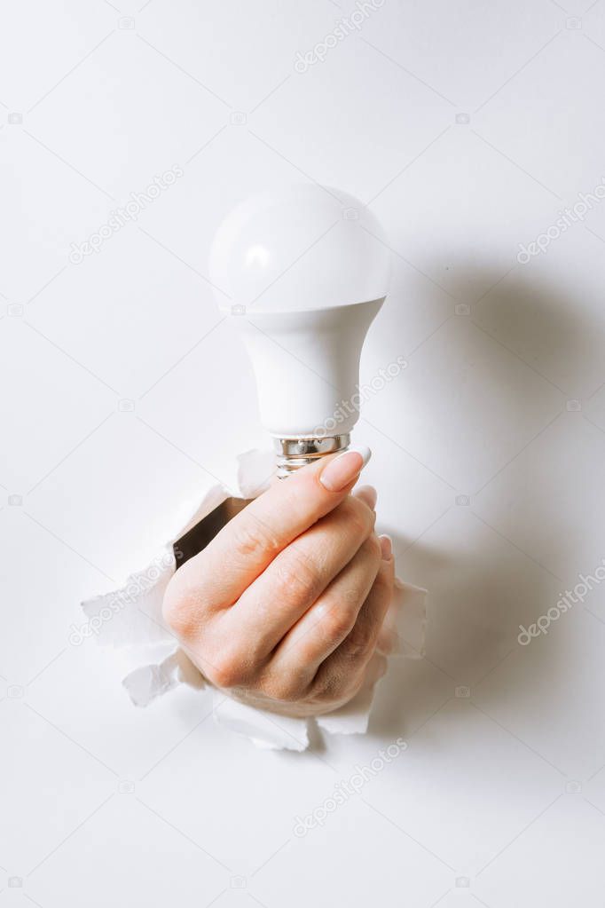Hand holding a light bulb breaking through the hole in white bac