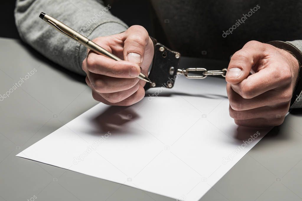 Hands of the criminal in handcuffs write a handle on paper. Sinc