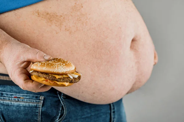 Grubas z hamburgerem w ręku na szarym tle. — Zdjęcie stockowe