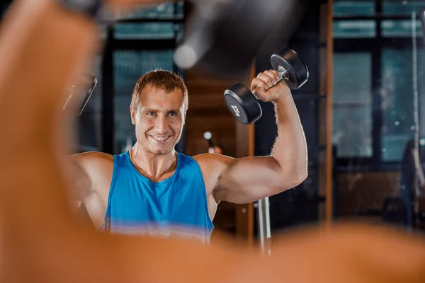 A young muscular guy does hard exercises with dumbbells for the rear deltas of the shoulders on a training bench in the gym.