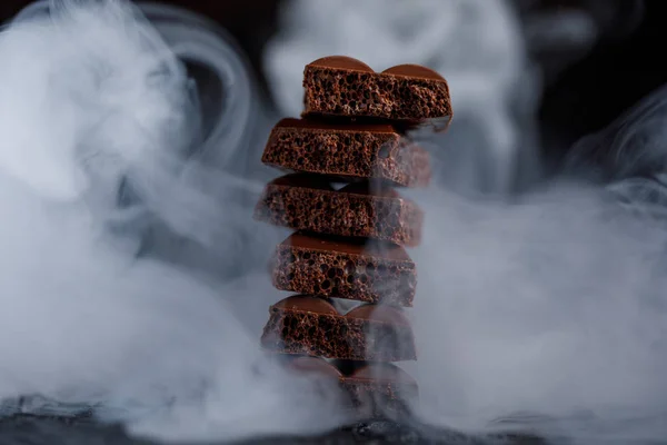 Chocolate poroso apilado sobre fondo negro. Pedazos de chocolate con leche, alineados en una torre, cubiertos de humo . — Foto de Stock