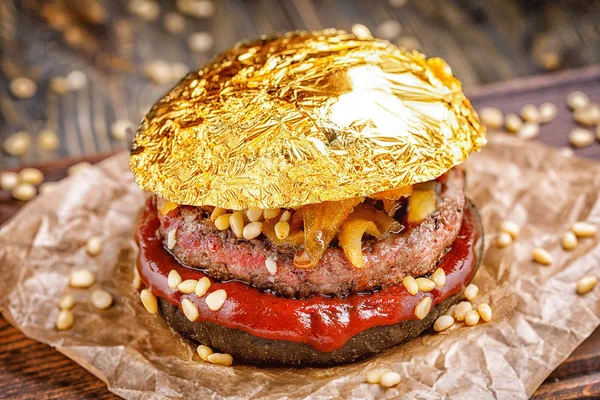 Goldener Rindfleisch-Burger mit Schwarzbrötchen, mit Rucola und Käse und Ketchup bestreut mit Pinienkernen, serviert auf braunem Papier auf einem rustikalen Holztisch vor dunklem Hintergrund. leckerer Goldburger — Stockfoto