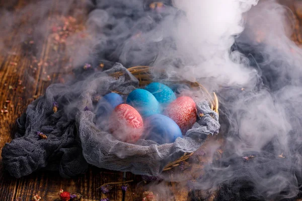 Cesta de mimbre con huevos inusualmente teñidos sobre un fondo de madera en humo . — Foto de Stock