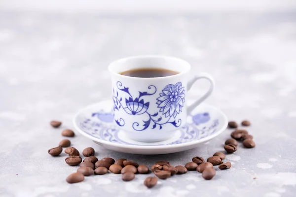 A beautiful porcelain coffee cup with hot black coffee. Coffee beans scattered around the background. Vintage china. A white-blue cup on a gray background. — Stock Photo, Image