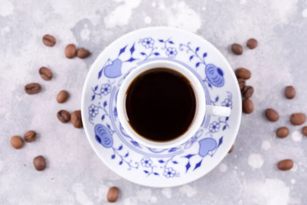 A beautiful porcelain coffee cup with hot black coffee. Coffee beans scattered around the background. Vintage china. A white-blue cup on a gray background. — Stock Photo, Image