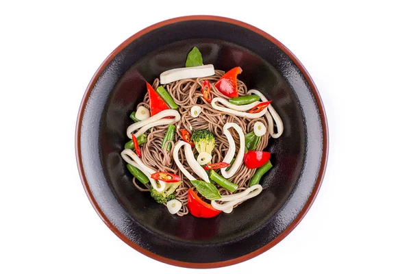 Fideos de trigo sarraceno con calamares y verduras con salsa teriyaki aislados sobre un fondo blanco. Fideos de trigo sarraceno en un plato negro . Imagen De Stock