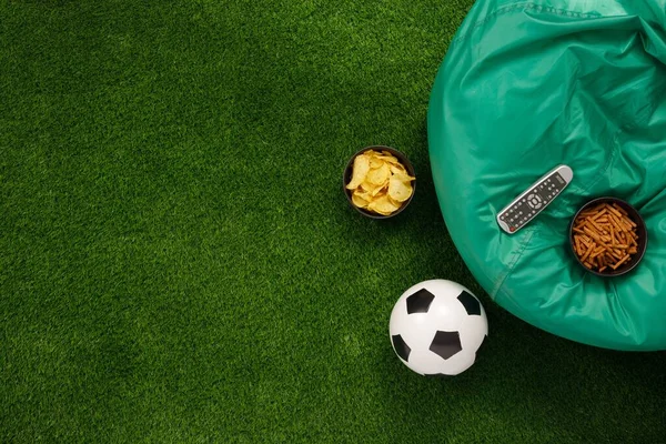 Soccer ball on a green field and ottoman for a fan with snacks and a TV remote control. The apartment was lying. The concept of football matches.