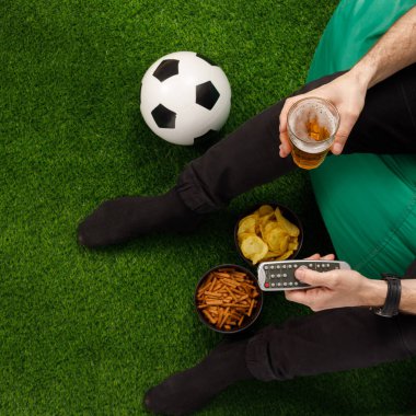 Bir adam TV 'de oturur ve futbol maçı izler. Sneki TV kumandası. Futbol topu. Yukarıdan bak. Uzayı kopyala Bir çimenliğin taklidi.