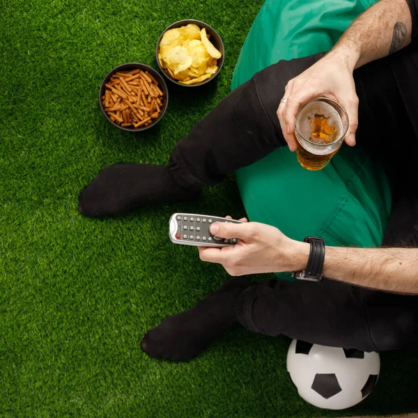 Ein Mann sitzt am Fernseher und schaut sich ein Fußballspiel an. Sneki TV Fernbedienung. Fußball. Blick von oben. Kopierraum. Nachahmung eines Rasens. — Stockfoto