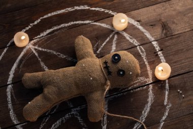 Tahta arka planda, dramatik ışıklandırması ve mumları olan bir vudu bebeği. Büyücülük, kara sanat ve okült kavramı. Çizilmiş bir yıldızın arka planındaki çuval bezi bebek. Oyuncak bebeğe iğneler saplanmış..