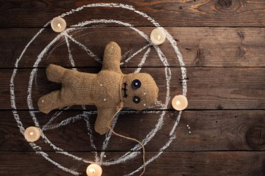 Tahta arka planda, dramatik ışıklandırması ve mumları olan bir vudu bebeği. Büyücülük, kara sanat ve okült kavramı. Çizilmiş bir yıldızın arka planındaki çuval bezi bebek. Oyuncak bebeğe iğneler saplanmış..