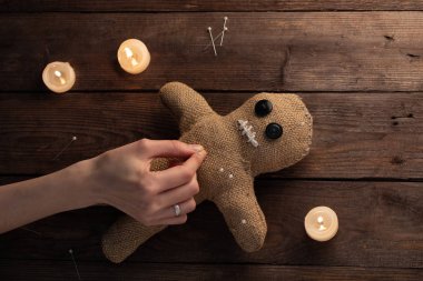 Voodoo doll on a wooden background with dramatic lighting and candles. The concept of witchcraft and black art. Burlap doll on a wooden background. Hands stick pins into a doll. Copy space.