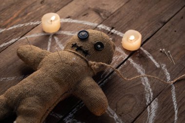 Tahta arka planda, dramatik ışıklandırması ve mumları olan bir vudu bebeği. Büyücülük, kara sanat ve okült kavramı. Çizilmiş bir yıldızın arka planındaki çuval bezi bebek. Oyuncak bebeğe iğneler saplanmış..