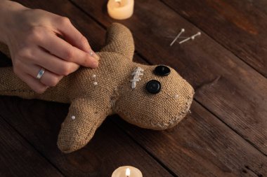 Voodoo doll on a wooden background with dramatic lighting and candles. The concept of witchcraft and black art. Burlap doll on a wooden background. Hands stick pins into a doll. Copy space.