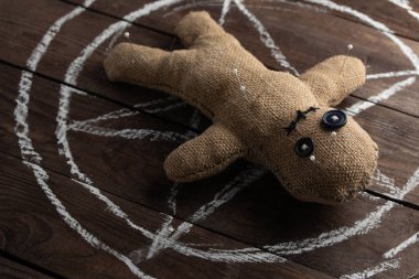 Tahta arka planda dramatik ışıklandırmalı bir vudu bebeği. Büyücülük, kara sanat ve okült kavramı. Çizilmiş bir yıldızın arka planındaki çuval bezi bebek. Oyuncak bebeğe iğneler saplanmış..