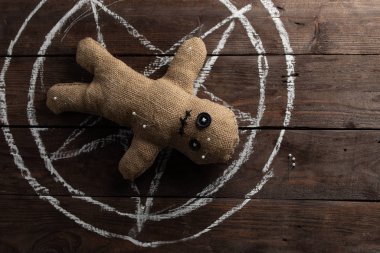 Tahta arka planda dramatik ışıklandırmalı bir vudu bebeği. Büyücülük, kara sanat ve okült kavramı. Çizilmiş bir yıldızın arka planındaki çuval bezi bebek. Oyuncak bebeğe iğneler saplanmış..
