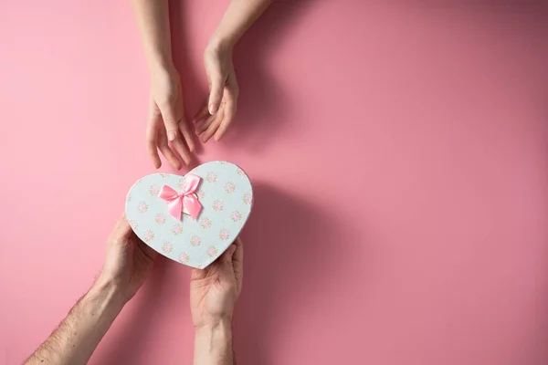 Valentine's Day celebration concept. A nice gift from a loved one. Box with a bow hands of a man and a woman on a delicate pink background. Copy space. Flat lay. — 스톡 사진