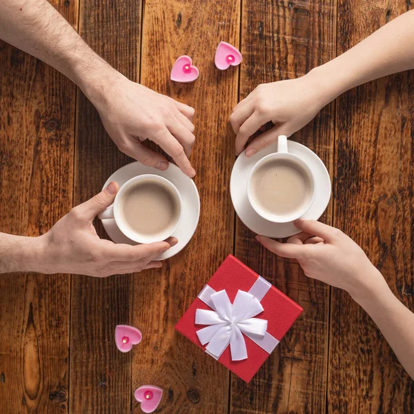 Valentine's Day celebration concept. A nice gift for your loved one. Hands of man and woman with coffee mugs on a wooden table background. Copy space. Flat lay. Close-up. — 스톡 사진