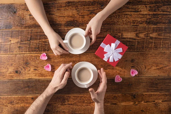 Valentine's Day celebration concept. A nice gift for your loved one. Hands of man and woman with coffee mugs on a wooden table background. Copy space. Flat lay. Close-up. — 스톡 사진